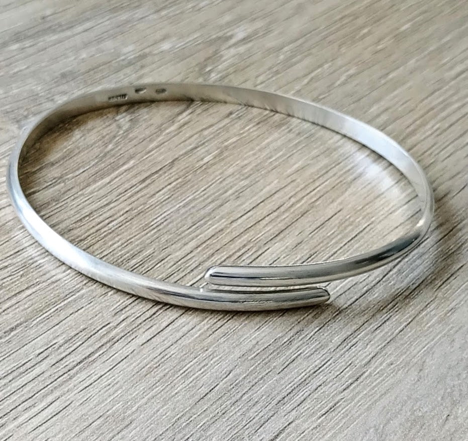 A polished silver open cuff bangle bracelet resting on a light gray wood-grain surface
