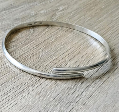 A polished silver open cuff bangle bracelet resting on a light gray wood-grain surface