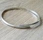 A polished silver open cuff bangle bracelet resting on a light gray wood-grain surface
